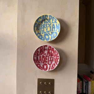May include: Two round ceramic bowls mounted on a wall. The top bowl is yellow with blue text reading CASPER AUBREY WILLIAM BOYD. The bottom bowl is white with red text reading HEBE ROSE MADELEINE BOYD. Both bowls feature hand-painted lettering.