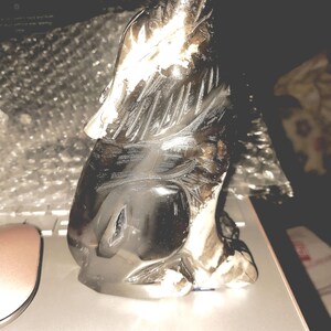 Puede incluir: Figura de lobo gris oscuro tallada. El lobo está representado en posición sentada, con la cabeza levantada y la boca abierta. La escultura tiene detalles de pelaje y rasgos faciales, y está hecha de un material reflectante.