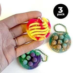 Può includere: Tre portachiavi colorati in resina. Uno è rosso e giallo con un design a onde, gli altri sono verde acqua e viola con piccole sfere. Ogni portachiavi ha una catena di metallo e il testo "3 pack" è visibile.