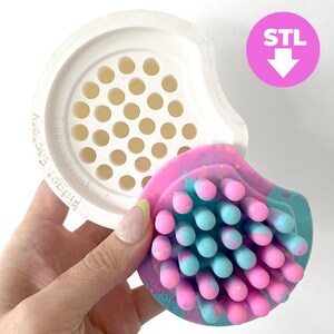Puede incluir: Un juguete fidget multicolor con detalles en rosa, azul y blanco. El juguete tiene una superficie texturizada con protuberancias redondeadas y se sostiene cerca de un componente blanco perforado. La imagen incluye el texto "STL" en un círculo rosa.