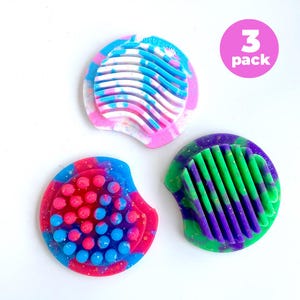May include: Three colorful fidget toys in a variety of designs. One is pink, white, and blue with a wavy pattern. Another is red, blue, and pink with small spheres. The third is green and purple with a striped design. The text "3 pack" is in a pink circle.