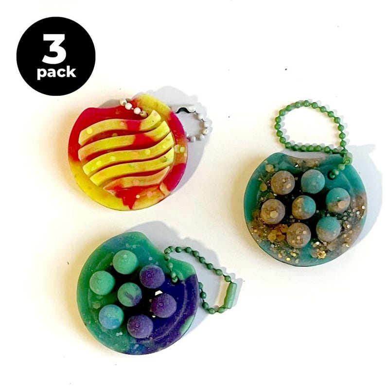 May include: Three colorful, round keychains. One is red and yellow with a striped design, another is teal with small brown spheres, and the third is green and purple with small purple spheres. The keychains have metal ball chains and the text "3 pack".