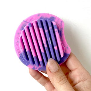 May include: A pink and purple fidget toy with a ridged centre and a rounded edge. The toy has the words "Fidget Factory" printed on the side. The ridges alternate between pink and purple.