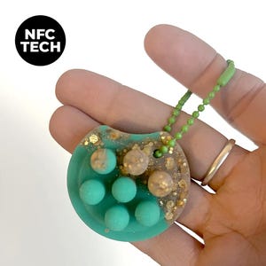 May include: A teal and gold NFC Tech keychain. The keychain has a teal base with six teal spheres and gold glitter accents. The top portion is a mix of gold glitter and beige spheres. The keychain is attached to a green ball chain.