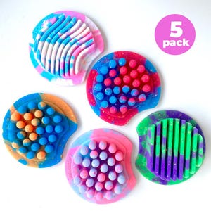 May include: Five brightly coloured, round silicone products with textured surfaces. Each has a unique design, including wavy lines and small, raised dots. The colour schemes include pink, blue, orange, green, and purple. A pink circle with the text "5 pack" is in the upper right.