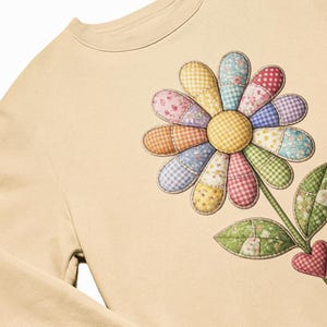 Peut inclure: Un sweat-shirt beige orné d'un grand motif floral. La fleur présente des pétales en patchwork aux motifs et couleurs variés, avec un centre en vichy jaune. Une tige et des feuilles vertes, ainsi qu'un cœur rouge, complètent le graphisme floral.