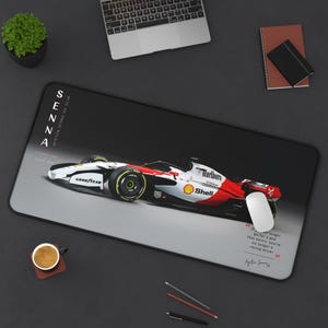 May include: A rectangular desk mat featuring a white, red, and black Formula 1 race car. The text reads SENNA AYRTON SENNA DA SILVA and includes a quote about racing drivers. A white computer mouse rests on the right side of the mat.