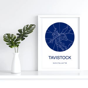 May include: A framed print displaying a navy blue circular map of Tavistock, with white lines representing streets and buildings. The town's name and coordinates are printed below. A white vase with green leaves is to the left.