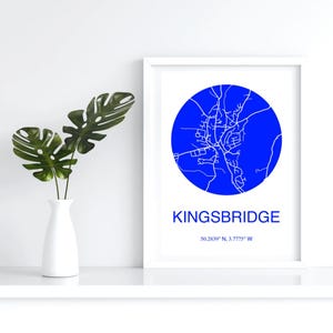 May include: A framed print featuring a blue circular map of Kingsbridge, with the town's name and coordinates below. The print is in a white frame, and a white vase with green leaves is to the left.