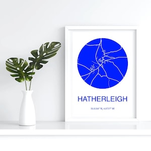 May include: A framed print featuring a blue circular map of Hatherleigh, with white lines representing streets. The town's name and coordinates are printed below the map. A white vase with green leaves is to the left.