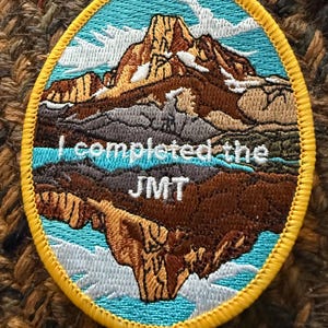 Peut inclure: Écusson brodé ovale avec un motif de paysage de montagne. L'écusson présente un ciel bleu, des montagnes brunes et le texte "I completed the JMT" en blanc. L'écusson a une bordure jaune.