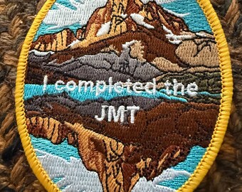 Może przedstawiać: Okrągła, haftowana naszywka z motywem górskiego krajobrazu. Naszywka przedstawia niebieskie niebo, brązowe góry i napis "I completed the JMT" w kolorze białym. Naszywka ma żółtą obwódkę.