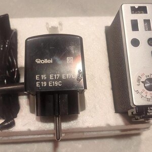 May include: A vintage Rollei camera light meter and charger set. The light meter is silver with a black leatherette grip and a dial. The charger is black with the Rollei logo and model numbers. The set is on a white foam base.