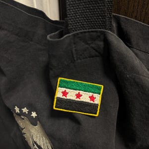 May include: Black tote bag with a patch of the Syrian flag. The patch has a green stripe, a white stripe, and a black stripe, with three red stars. An embroidered eagle and stars are also visible on the bag.