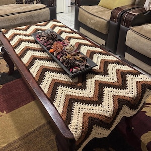 Puede incluir: Una mesa de centro de madera marrón con un camino de mesa de ganchillo. El camino de mesa presenta un patrón de chevron en crema, marrón claro y marrón oscuro. Una bandeja rectangular negra con objetos decorativos está sobre la mesa.