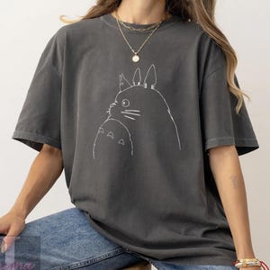 Op de afbeelding: Donkergrijs T-shirt met een witte omtrekillustratie van twee Totoro-personages. Het T-shirt heeft een losse pasvorm en korte mouwen. Het ontwerp is een eenvoudige lijntekening van de iconische animatiefiguren.