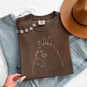 Op de afbeelding: Een bruin T-shirt met een witte omtrekillustratie van Totoro uit de animatiefilm My Neighbor Totoro. Het shirt wordt gecombineerd met een lichtblauwe jeans, een bruine hoed en een bloemenketting.