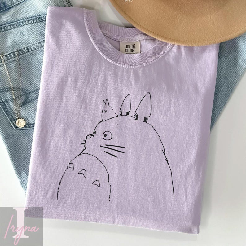 Op de afbeelding: Een lichtpaars T-shirt met een zwarte lijntekening van een Totoro-personage. Het T-shirt heeft een ronde hals en een Comfort Colors-label. Het shirt is opgevouwen op een blauwe spijkerbroek en een bruine hoed.