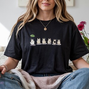 Op de afbeelding: Zwart T-shirt met een rij cartoonfiguren. De figuren hebben witte lichamen, grijze buiken en verschillende gezichtsuitdrukkingen. Boven het eerste personage staat een groen blad. Het T-shirt wordt gedragen.