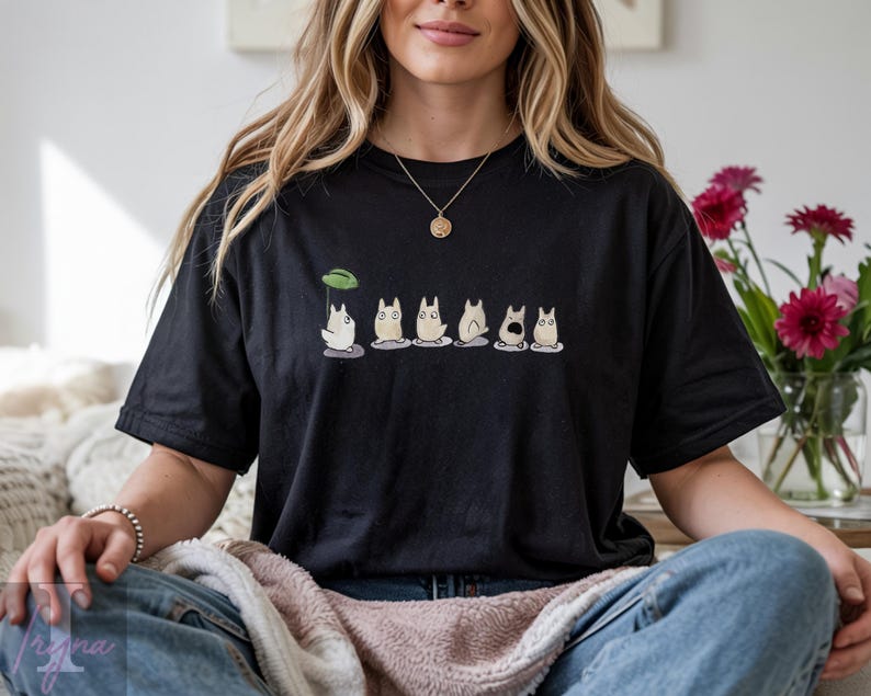Op de afbeelding: Zwart T-shirt met een rij cartoonfiguren. De figuren hebben witte lichamen, grijze buiken en verschillende gezichtsuitdrukkingen. Boven het eerste personage staat een groen blad. Het T-shirt wordt gedragen.