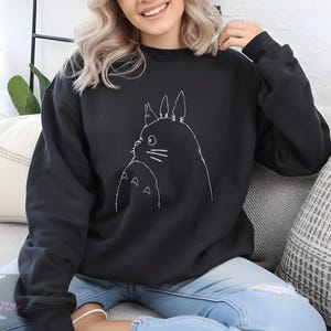 Op de afbeelding: Zwart sweatshirt met ronde hals en een witte lijntekening van een stripfiguur. Het ontwerp toont een groot, rond wezen met puntige oren en een kleiner wezen eronder. Het sweatshirt is effen zwart van kleur.