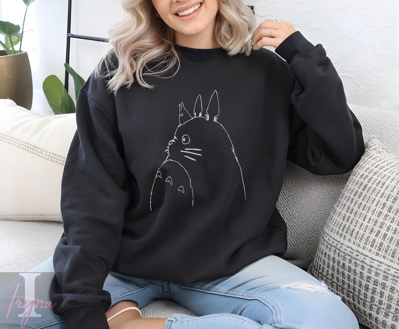 Op de afbeelding: Zwart sweatshirt met ronde hals en een witte lijntekening van een stripfiguur. Het ontwerp toont een groot, rond wezen met puntige oren en een kleiner wezen eronder. Het sweatshirt is effen zwart van kleur.