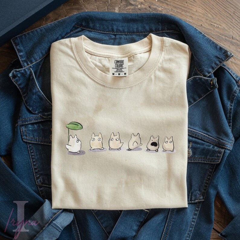 Op de afbeelding: Cr&egrave;mekleurig T-shirt met een rij cartoonfiguren onder een groen blad. De figuren hebben ronde lichamen en verschillende uitdrukkingen. Het T-shirt ligt op een spijkerjasje.