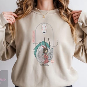 May include: Beige sweatshirt featuring a graphic from the animated film Spirited Away. The design includes a white mask, a dragon, and a girl in a red dress. Japanese text is also included in the design.