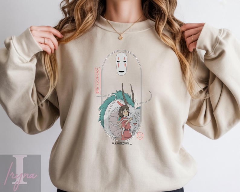 May include: Beige sweatshirt featuring a graphic from the animated film Spirited Away. The design includes a white mask, a dragon, and a girl in a red dress. Japanese text is also included in the design.