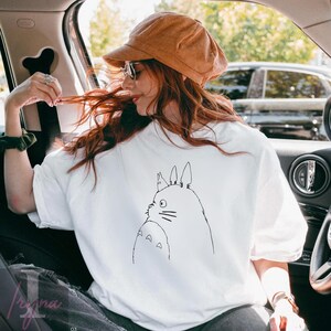 Op de afbeelding: Wit sweatshirt met korte mouwen met een zwarte lijntekening van een stripfiguur. Het sweatshirt wordt gedragen door een persoon in een auto, met een bruine corduroy pet en zonnebril.