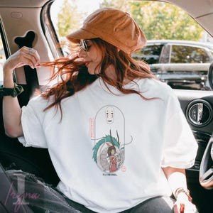 May include: White t-shirt featuring a Japanese anime-style illustration. The design includes a dragon, a girl in a red dress, and a faceless spirit. Japanese text is also present. The shirt is worn by a person in a car.