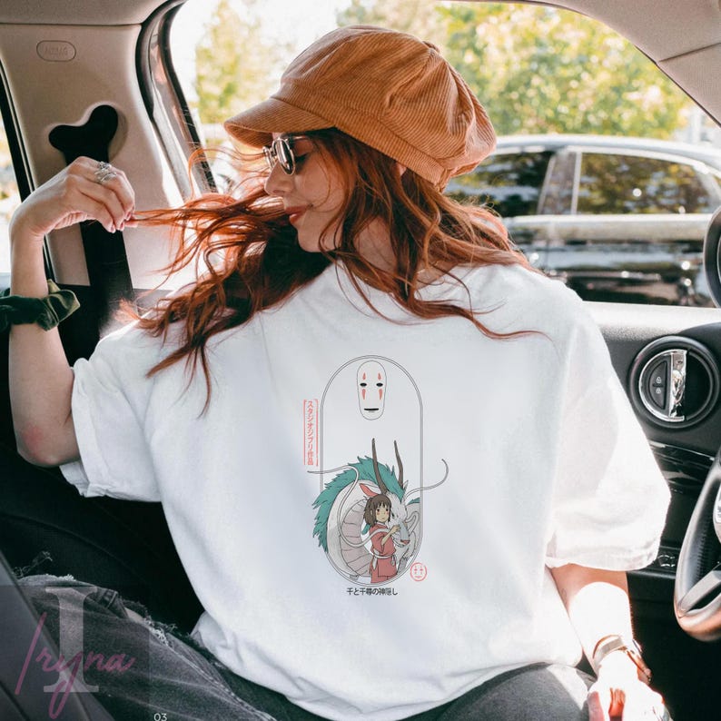 May include: White t-shirt featuring a Japanese anime-style illustration. The design includes a dragon, a girl in a red dress, and a faceless spirit. Japanese text is also present. The shirt is worn by a person in a car.