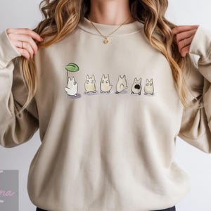 Op de afbeelding: Beige sweatshirt met een aquarel illustratie van cartoonfiguren. Het ontwerp toont een wit personage met een groene bladeren paraplu, gevolgd door andere personages in verschillende poses. De sweatshirt heeft een ronde hals.
