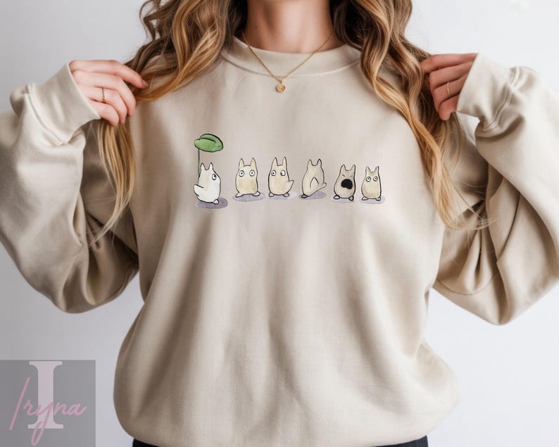 Op de afbeelding: Beige sweatshirt met een aquarel illustratie van cartoonfiguren. Het ontwerp toont een wit personage met een groene bladeren paraplu, gevolgd door andere personages in verschillende poses. De sweatshirt heeft een ronde hals.