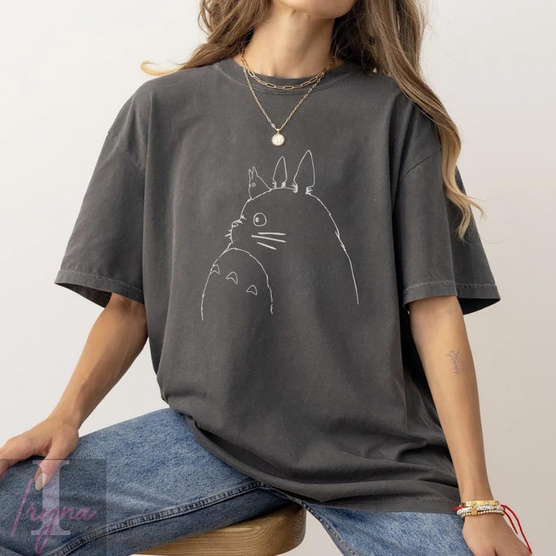 Op de afbeelding: Donkergrijs T-shirt met een witte omtrekillustratie van twee Totoro-personages. Het T-shirt heeft een losse pasvorm en korte mouwen. Het ontwerp is een eenvoudige lijntekening van de iconische animatiefiguren.