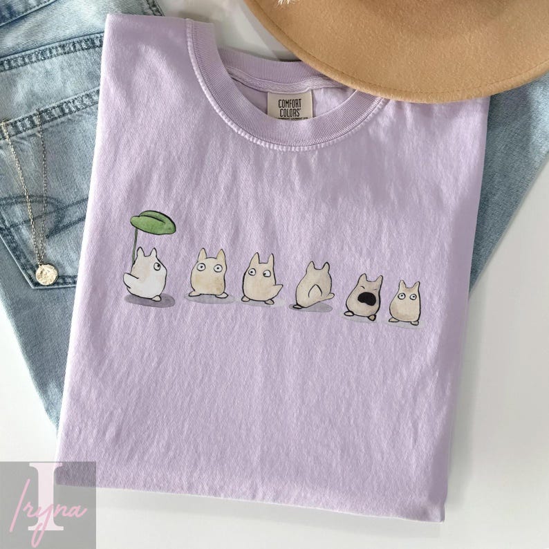 Op de afbeelding: Lichtpaars T-shirt met een reeks cartoonfiguren. E&eacute;n figuur houdt een groene paraplu vast. Het T-shirt is gemaakt van een zacht materiaal en heeft een Comfort Colors label.