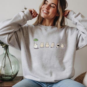 Op de afbeelding: Een lichtgrijs sweatshirt met een ronde hals en lange mouwen. Het heeft een reeks cartoonfiguren, waarvan &eacute;&eacute;n een paraplu van bladeren vasthoudt. De figuren staan in verschillende poses.