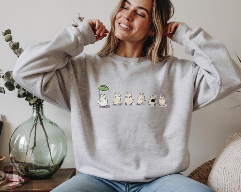 Op de afbeelding: Een lichtgrijs sweatshirt met een ronde hals en lange mouwen. Het heeft een reeks cartoonfiguren, waarvan &eacute;&eacute;n een paraplu van bladeren vasthoudt. De figuren staan in verschillende poses.