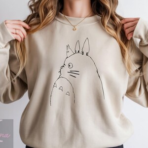 Op de afbeelding: Beige sweatshirt met een zwarte lijntekening van Totoro uit de animatiefilm My Neighbor Totoro. De illustratie toont het zijaanzicht van Totoro met een kleiner wezen op zijn hoofd. De sweatshirt heeft een ronde hals en lange mouwen.