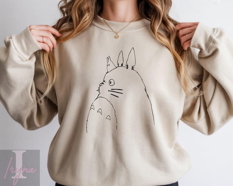 Op de afbeelding: Beige sweatshirt met een zwarte lijntekening van Totoro uit de animatiefilm My Neighbor Totoro. De illustratie toont het zijaanzicht van Totoro met een kleiner wezen op zijn hoofd. De sweatshirt heeft een ronde hals en lange mouwen.