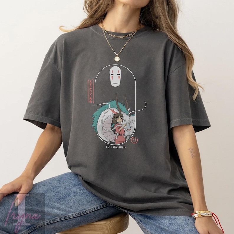 May include: Dark gray t-shirt featuring a graphic from the animated film Spirited Away. The design includes the character No-Face, a girl in a pink dress, and a dragon. Japanese text is also included in the design.
