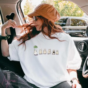Op de afbeelding: Wit sweatshirt met ronde hals en een cartoonontwerp. Het ontwerp toont een wit personage met een groene bladeren paraplu, gevolgd door verschillende bruinachtige personages met verschillende uitdrukkingen. De trui is een casual stijl.