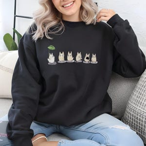 Op de afbeelding: Zwarte sweatshirt met een cartoonontwerp van verschillende witte, katachtige wezens. E&eacute;n wezen houdt een groene bladeren paraplu vast. Het ontwerp is gecentreerd op de borst. Het sweatshirt is gemaakt van een zacht materiaal.