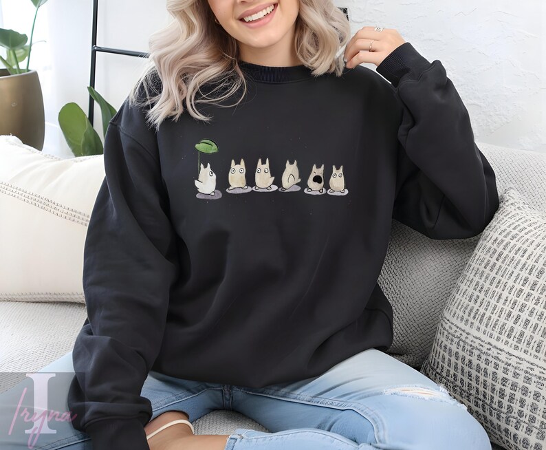 Op de afbeelding: Zwarte sweatshirt met een cartoonontwerp van verschillende witte, katachtige wezens. E&eacute;n wezen houdt een groene bladeren paraplu vast. Het ontwerp is gecentreerd op de borst. Het sweatshirt is gemaakt van een zacht materiaal.