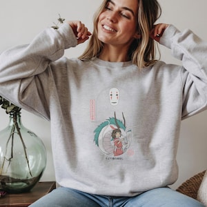 May include: A light gray sweatshirt featuring a graphic from the animated film Spirited Away. The design includes the character No-Face, a dragon, and a girl in a red dress. Japanese text is also included in the design.
