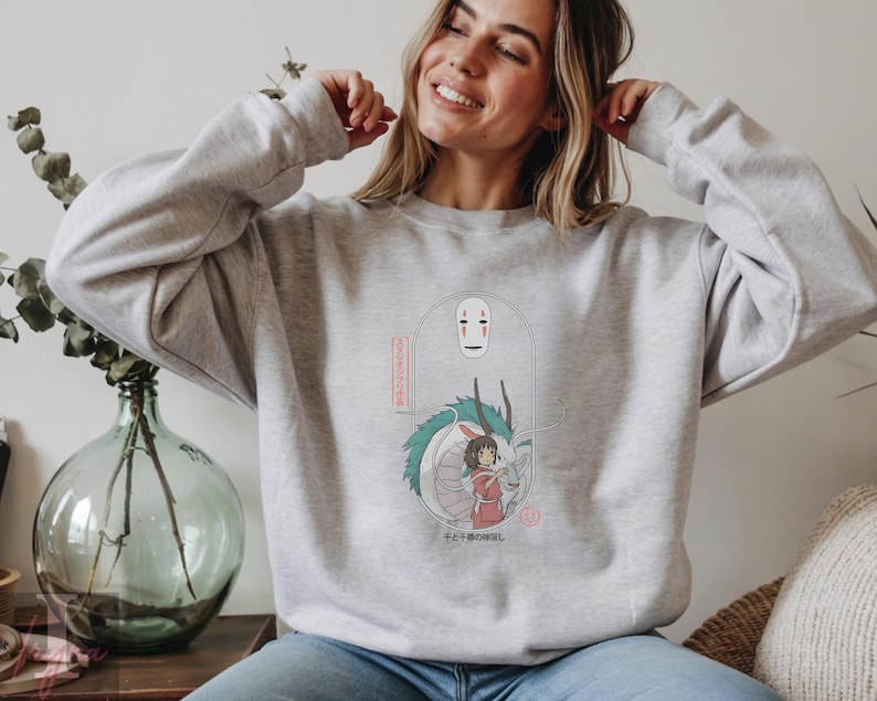 May include: A light gray sweatshirt featuring a graphic from the animated film Spirited Away. The design includes the character No-Face, a dragon, and a girl in a red dress. Japanese text is also included in the design.