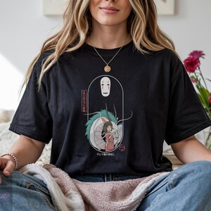May include: Black t-shirt featuring a graphic from the animated film Spirited Away. The design includes the character No-Face, a dragon, and a girl in a red dress. Japanese text is also included in the design.