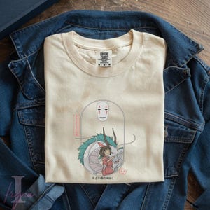 May include: Cream-colored t-shirt featuring a graphic from the animated film, Spirited Away. The design includes a white dragon, a girl in a red dress, and the faceless No-Face character. Japanese text is also included.