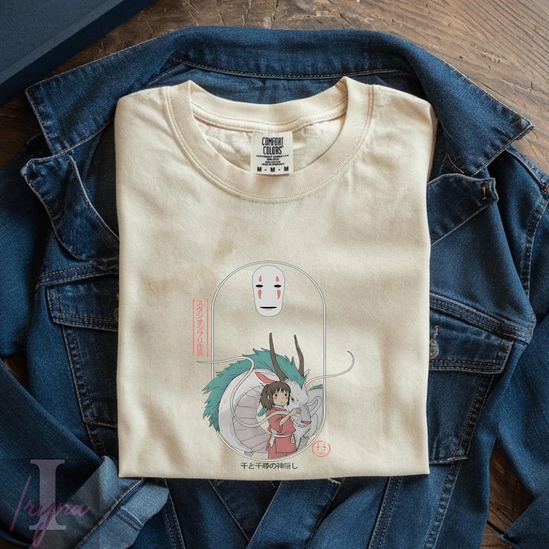 May include: Cream-colored t-shirt featuring a graphic from the animated film, Spirited Away. The design includes a white dragon, a girl in a red dress, and the faceless No-Face character. Japanese text is also included.