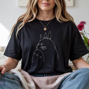 Op de afbeelding: Zwart T-shirt met een witte omtrek van twee Totoro-personages. Het ontwerp is gecentreerd op de voorkant. Het shirt is gemaakt van een zacht materiaal en heeft korte mouwen.